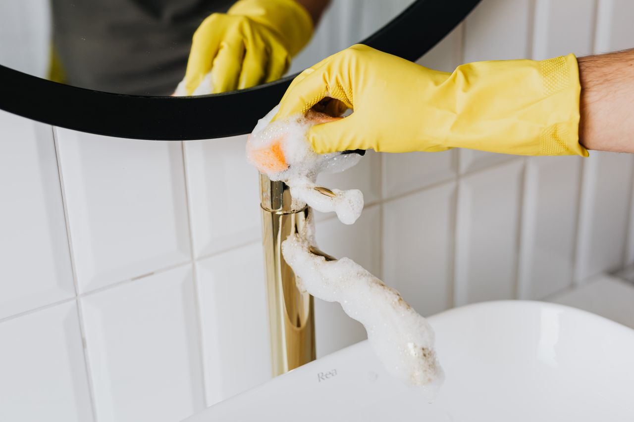 services-04 Anonymous person wearing yellow latex gloves washing golden faucet with sponge in modern bathroom while cleaning house